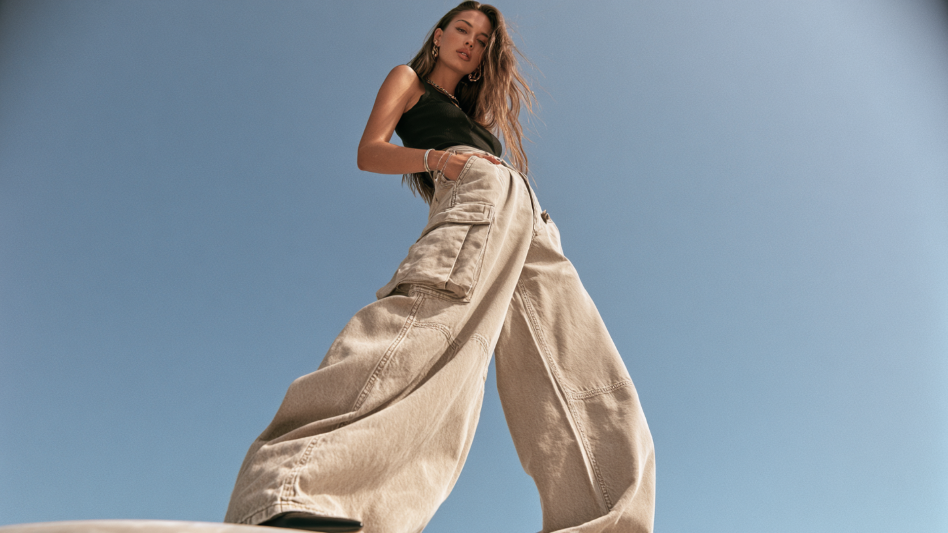 Woman wearing oversized beige cargo pants with black sleeveless top under blue sky fashion editorial style full body pose