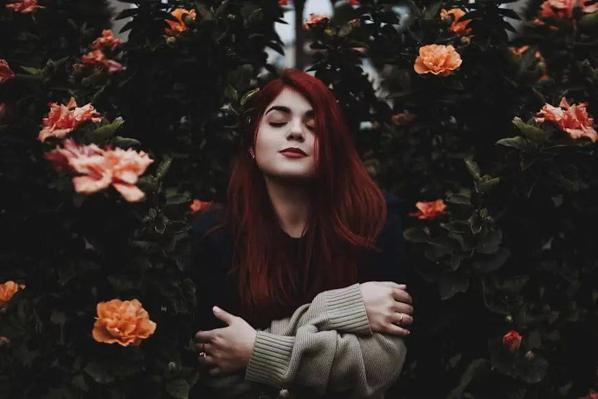 Woman with red hair embracing herself among vibrant orange flowers in a garden setting